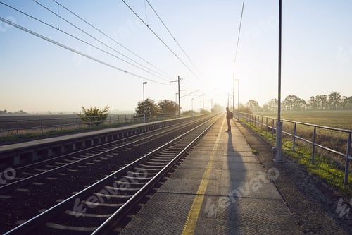 Preview: Alone young man with backpack waiting for train at railroad station at sunrise.