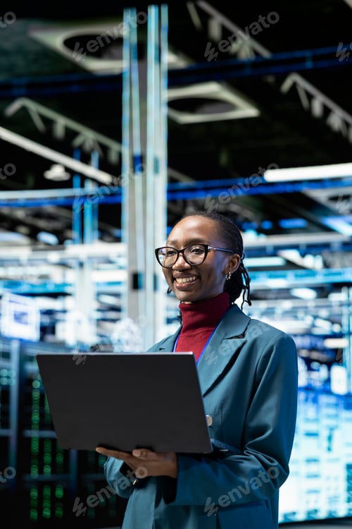 Preview: Cheerful female technician in data center using laptop to monitor rigs metrics