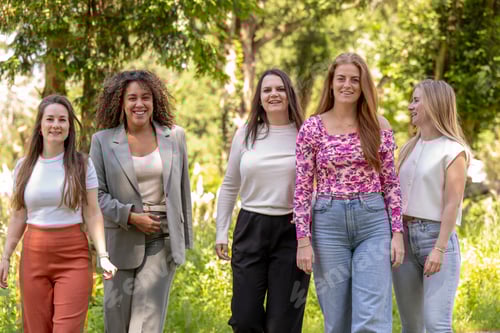 Preview: Five women smiling and walking outdoors in a park-like setting during the daytime.