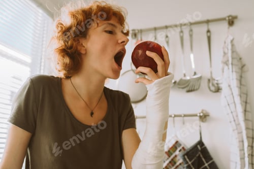 Preview: teenage girl with broken arm in cast in kitchen sitting on countertop eating apple