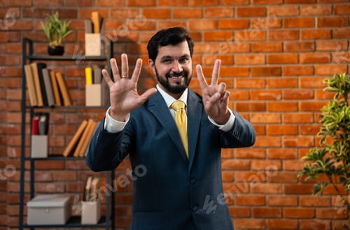 Preview: Corporate Indian man counting digits with fingers while standing in office against brick wall
