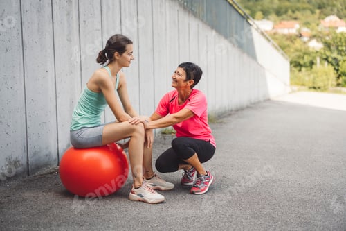 Preview: A fitness coach encourages a young woman sitting on a stability ball. They are outdoors, with a conc