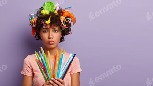 Preview: Discontent Afro American female volunteer holds colorful disposable plastic drinking straws, has gar