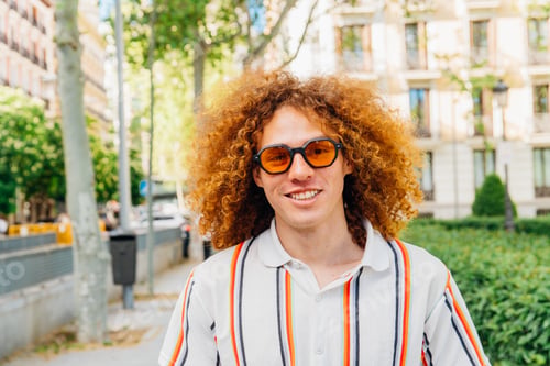 Preview: Curly-haired male wearing sunglasses, beaming with confidence near urban greenery
