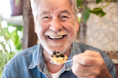 Preview: Close up on happy bearded senior man eating a fruit cake in coffee shop enjoying break or breakfast