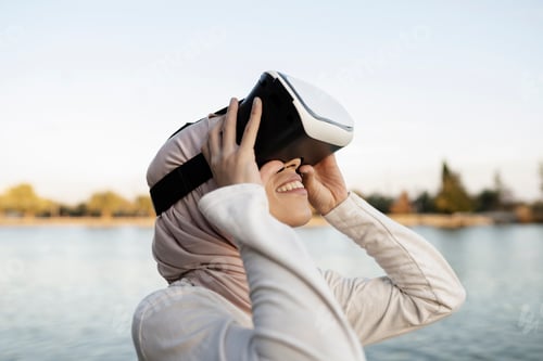 Preview: Side view of positive Arab female in traditional hijab and modern VR goggles looking up while