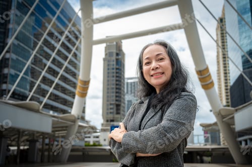 Preview: A confident mature Asian businesswoman stands in the big urban city with her arms crossed.
