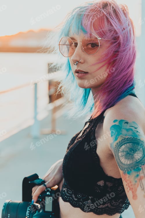 Preview: Young female with pink and blue hair and tattoos holding a camera standing near the sea