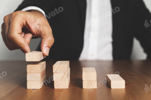 Preview: Hand of a person with a suit placing a wooden block on the wooden block tower