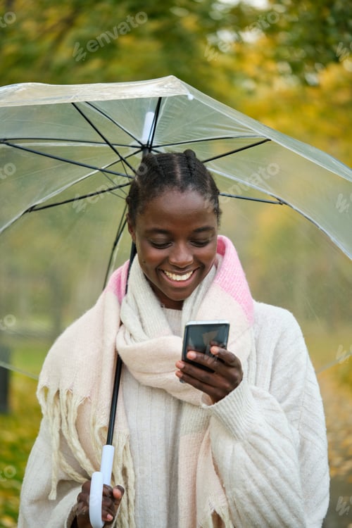 Preview: African woman with an umbrella smiling and using the phone in autumn.
