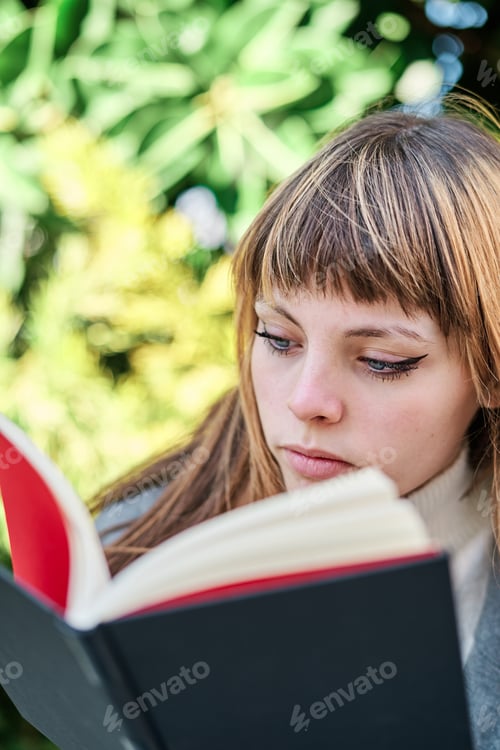 Preview: Close-up portrait of blue-eyed Caucasian blonde young woman reading a book in the park