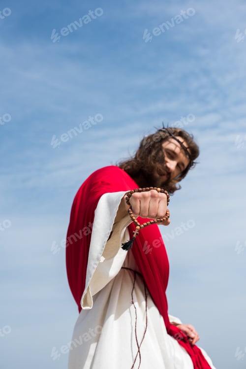 Preview: low angle view of Jesus in robe, red sash and crown of thorns showing fist with rosary against blue