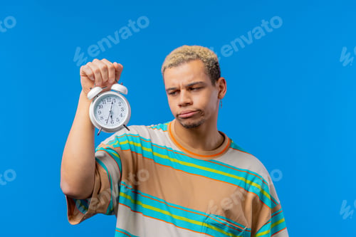 Preview: Woken up by alarm clock sleepy man holding it in hand. Blue background. Early 6 o'clock in morning