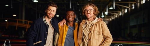 Preview: multicultural male friends smiling and standing near racing cars inside of karting track, banner