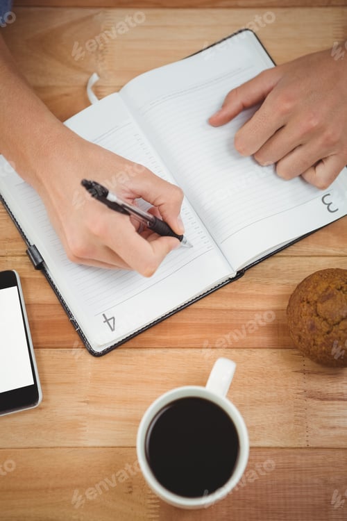 Preview: Cropped image of man writing on diary with muffin and coffee at desk in office