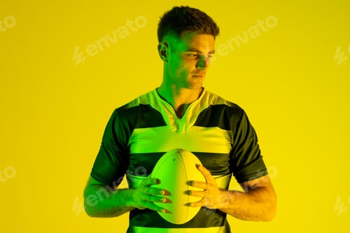 Preview: Caucasian male rugby player with rugby ball over yellow lighting