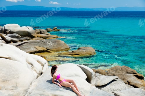 Preview: beautiful young woman sitting on the wonderful stone coast in greece in swimsuit