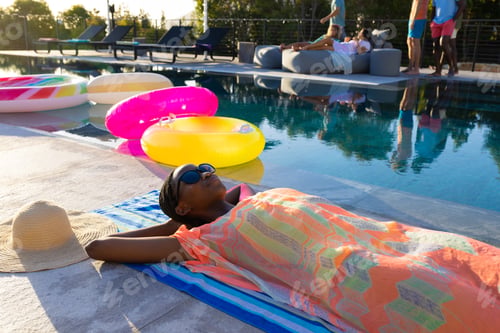 Preview: Happy african american woman lying on towel and sunbathing next to swimming pool in garden