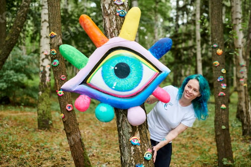 Preview: Smiling young adult woman with dyed turquoise hair in white T shirt near big eye art installation
