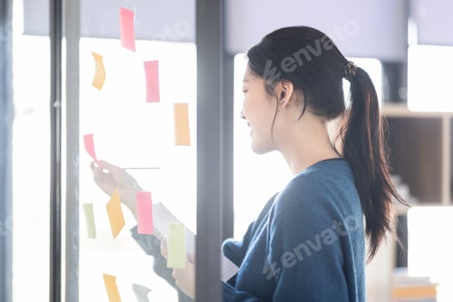Preview: Portrait shot of Cheerful young adult business Asian woman sticking adhesive paper notes to a glass