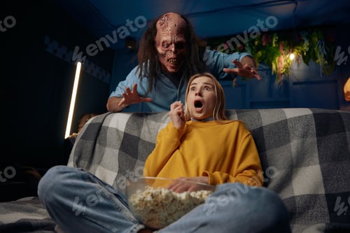 Preview: Shocked woman watching movie with popcorn. Man wearing zombie mask behind wants to scary her