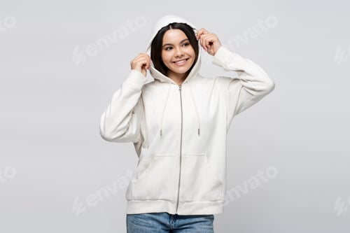 Preview: Smiling latin teenage girl wearing hood on head, standing isolated on gray background, looking away