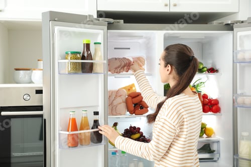 Preview: Young woman taking ham out of refrigerator in kitchen