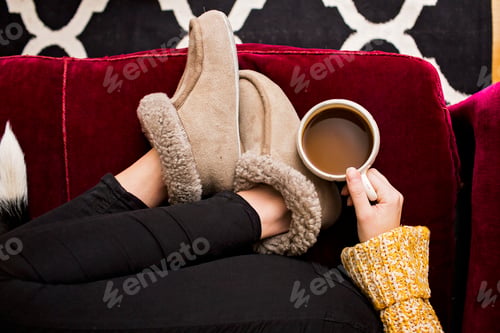 Preview: Low section of woman holding coffee cup while sitting on sofa at home