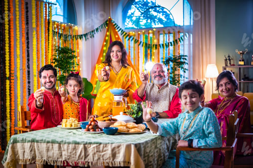 Preview: Portrait of happy Indian family in traditional outfit celebrating Diwali at home eating food