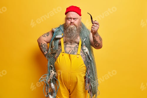 Preview: Angry irritated bearded boatswain looks with sullen expression directly at you holds smoking pipe dr