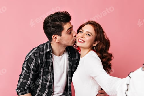 Preview: Brunette man in casual shirt kissing wife. Blissful couple making selfie on pink background.