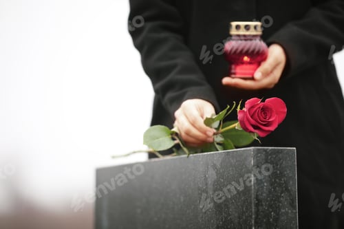 Preview: Woman with candle near black granite tombstone outdoors, focus on red rose. Funeral ceremony