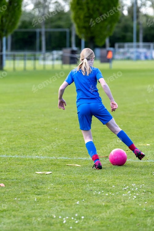 Preview: Female football player in blue uniform kicking a pink ball on a lush green field.