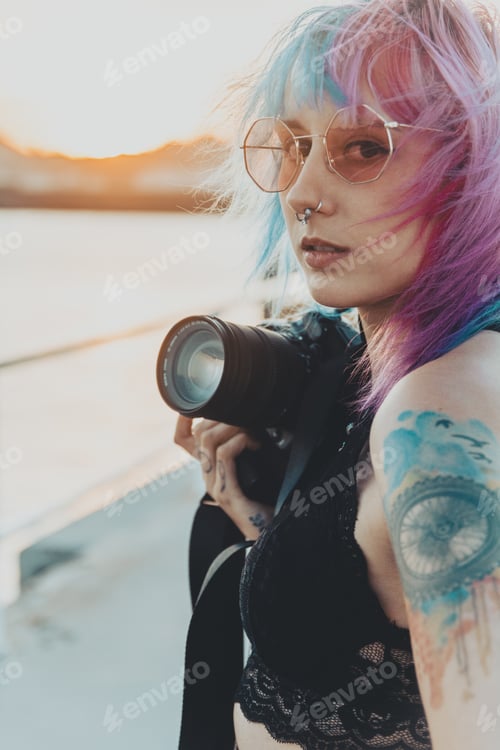 Preview: Young female with pink and blue hair and tattoos holding a camera standing near the sea