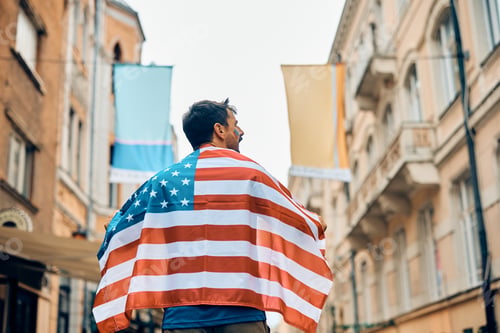 Preview: Rear view of sports fan covered with American national flag walking on the street.