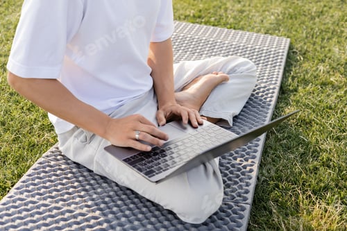 Preview: cropped view of man using laptop while sitting in easy pose on yoga mat on green grass