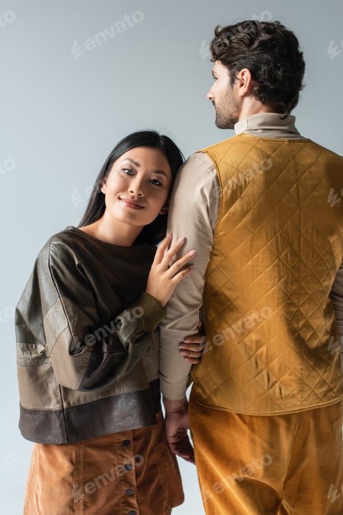 Preview: happy asian woman in leather pullover leaning on man in yellow vest isolated on grey