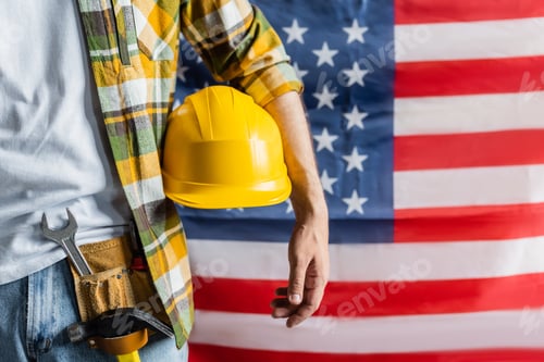 Preview: cropped view of workman in plaid shirt and tool belt holding hardhat near blurred usa flag, labor