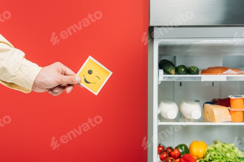 Preview: cropped view of man holding smiley face near open fridge with fresh food on shelves isolated on red