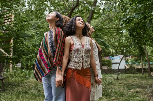 Preview: smiling and trendy interracial girlfriends holding hands and standing back to back in retreat center