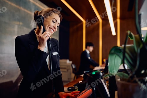 Preview: Happy female receptionist answering a phone call while working at hotel front desk.
