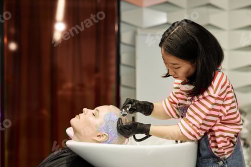 Preview: Young Asian Hairstylist Washing Hair of Young Woman in Salon Sink
