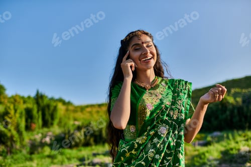 Preview: cheerful young indian woman touching face while posing in sari with landscape on background