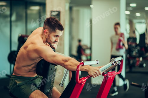 Preview: A shirtless sportsman in shape is reaching for a handle on an exercise machine in a gym.