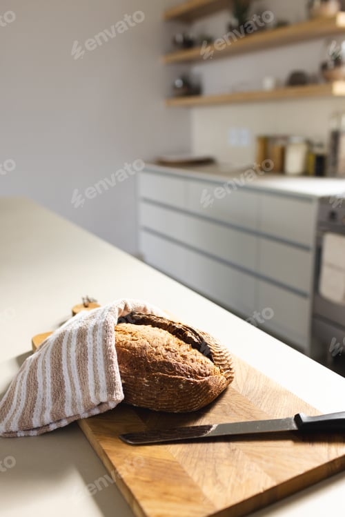 Preview: A loaf of bread rests on a wooden cutting board, partially covered by a striped towel, with copy spa