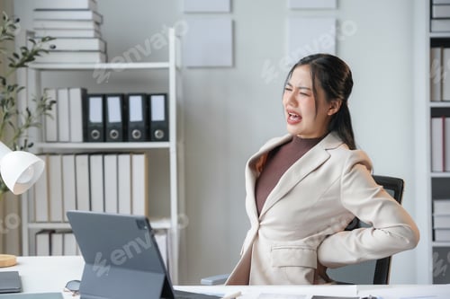 Preview: Tired office worker grimaces in pain, holding her lower back at her desk.