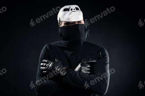 Preview: Thief in black balaclava and white mask holding gun in folded arms on black