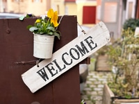 Preview: Word "Welcome" Written on Old Brown Rustic Wooden Background Decorated With Potted Flowering Plant