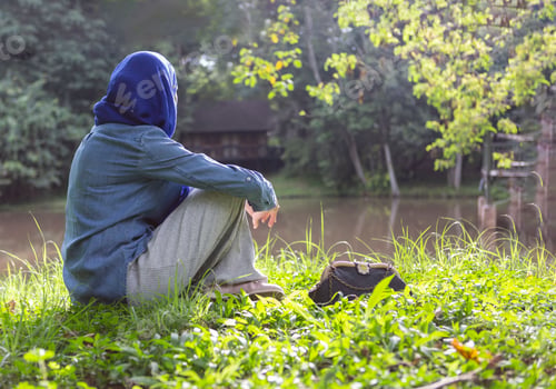 Preview: Person with a jacket and a hood sitting on green grass against a lake on a sunny day