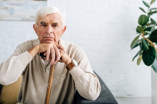 Preview: senior man looking at camera and holding wooden cane in apartment